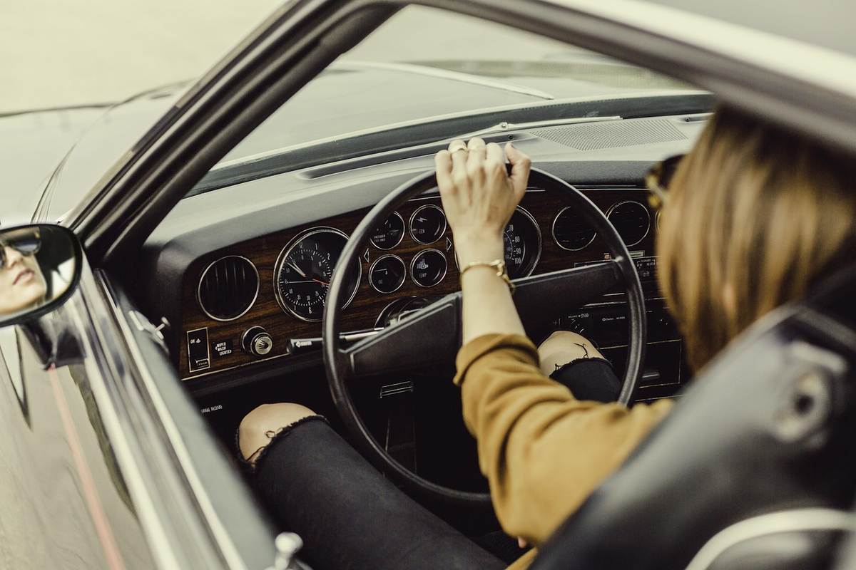 Une femme conduit une voiture ancienne, main sur le volant, regard vers le miroir